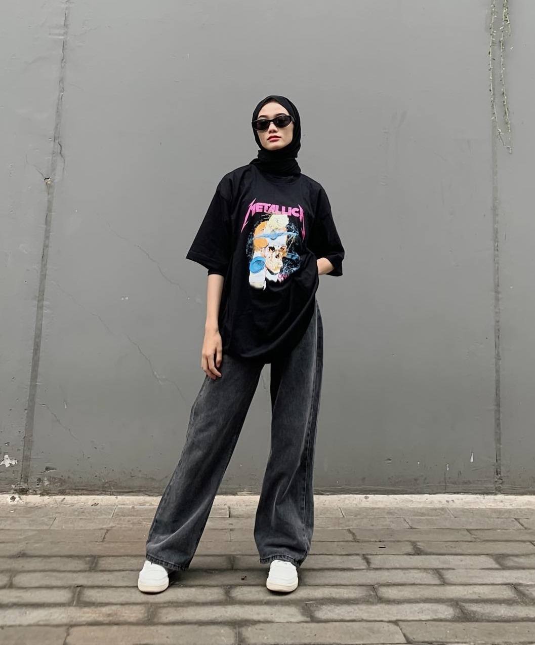 Wanita Indonesia memakai hijab hitam dengan kaos band Metallica dan celana jeans panjang.