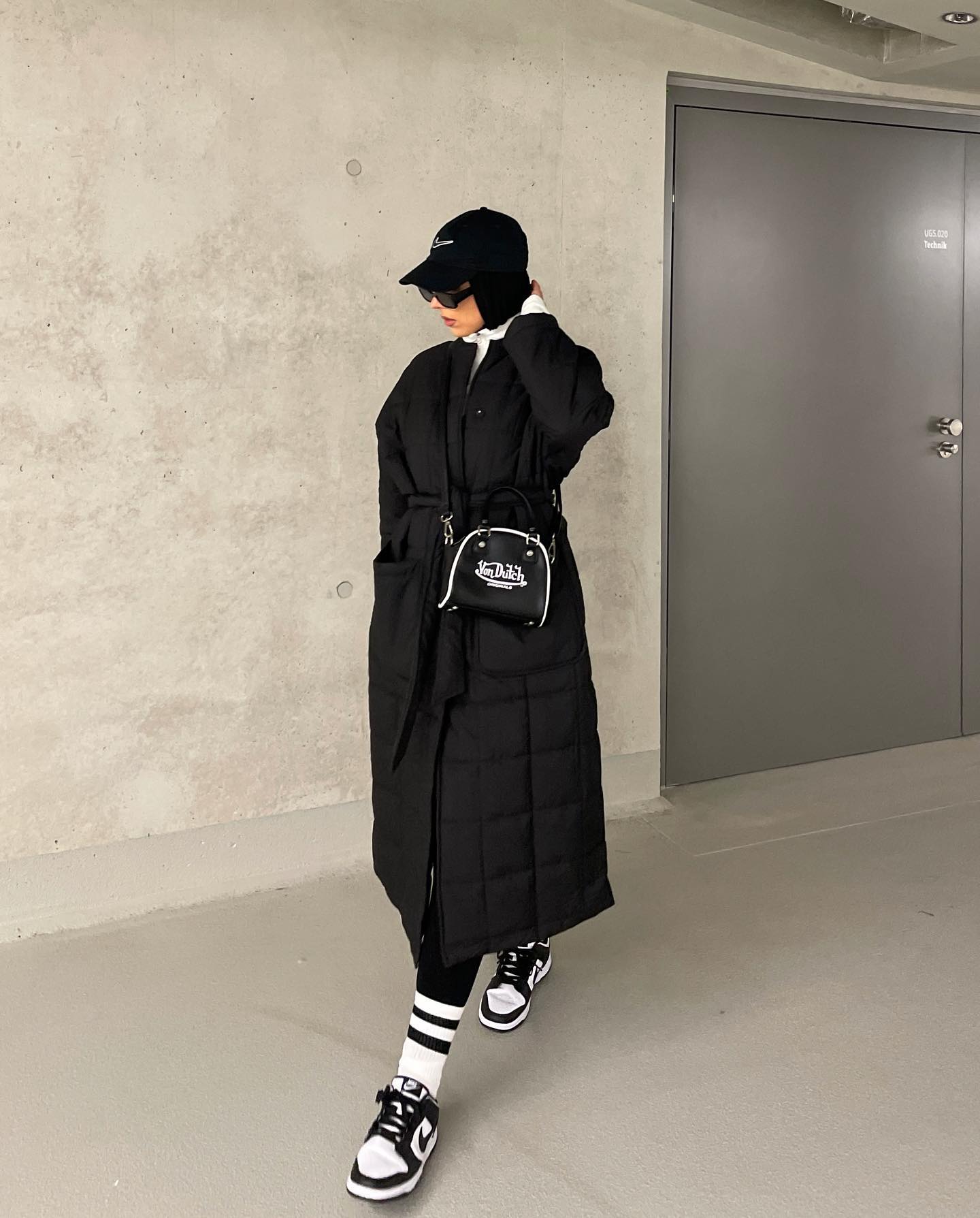 Wanita kaukasia mengenakan long coat hitam, legging hitam dan sepatu sneakers.