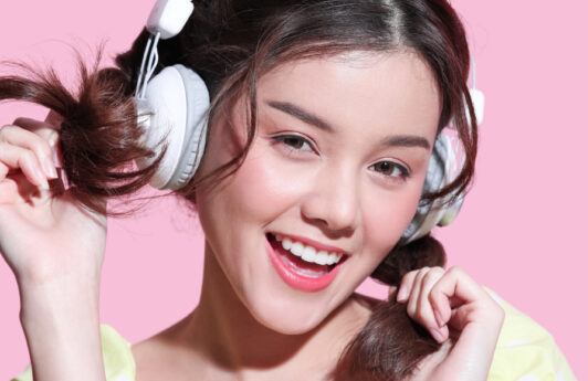 Wanita Asia menggunakan headphone warna putih sedang tertawa gembira dengan kedua tangan memegang rambut.
