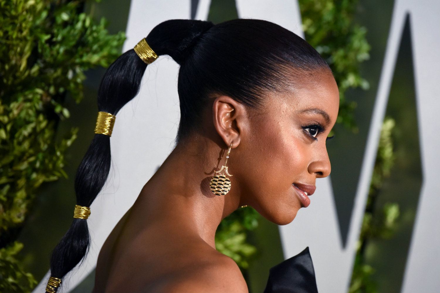 Vista de perfil de mujer afroamericana, lleva el cabello tirante hacia atrás atado en una bubbly pony tail con banditas en dorado 