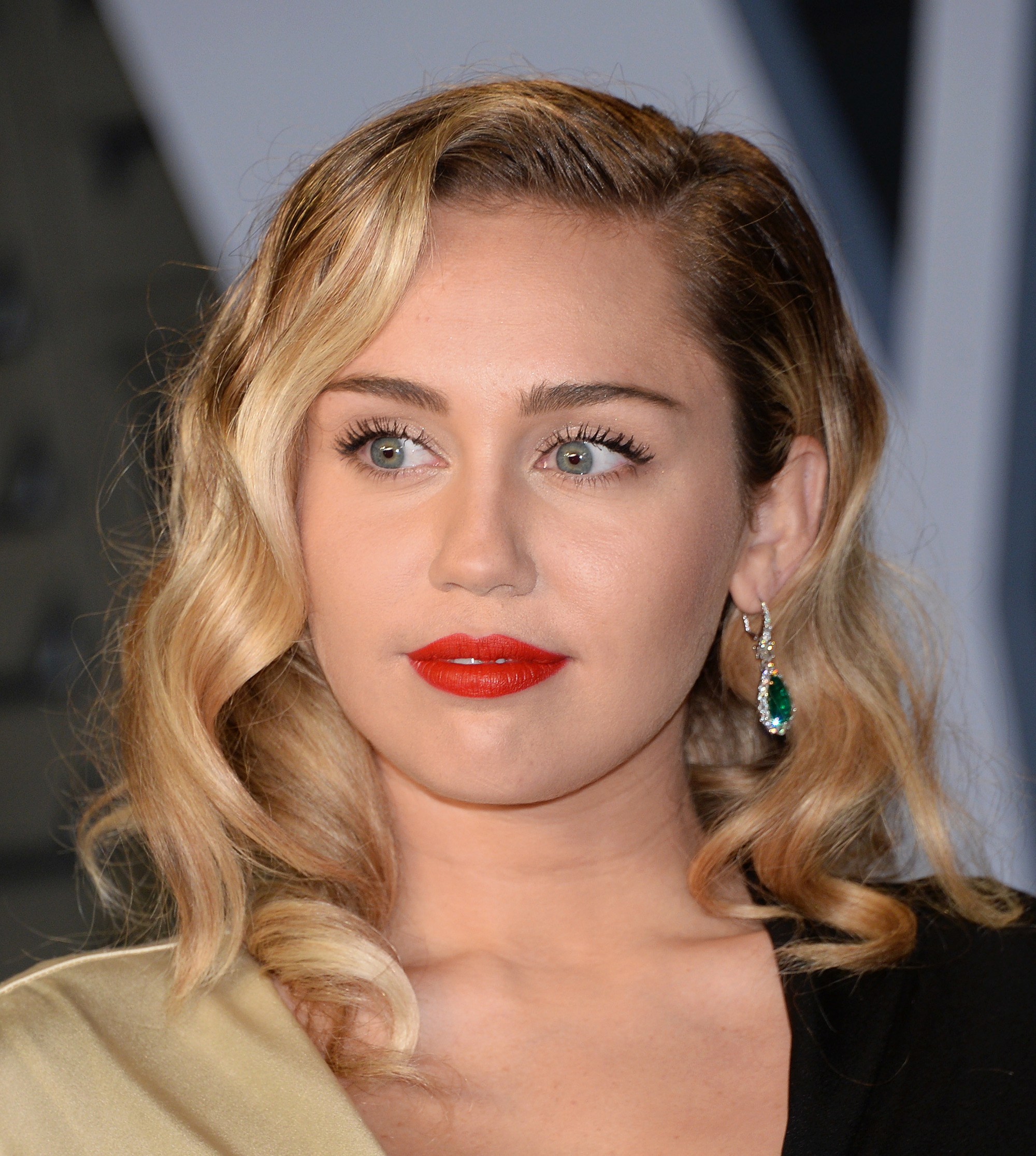 Miley Cyrus con cabello rubio por los hombros peinado con ondas y raya a un costado con un semi jopo