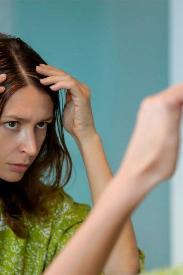 mujer joven frente al espejo en bata examinando su cuero cabelludo, exfoliar el cuero cabelludo