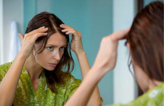 mujer joven frente al espejo en bata examinando su cuero cabelludo, exfoliar el cuero cabelludo