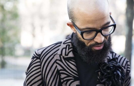 Man with shaved bald head and long dark brown beard wearing glasses and black strip blazer in street style image.
