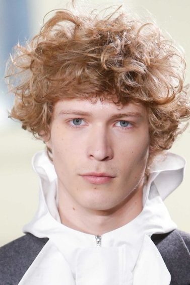 Curly hairstyles for men: Man on runway with blonde curly messy hair wearing a high neck ruffle shirt.