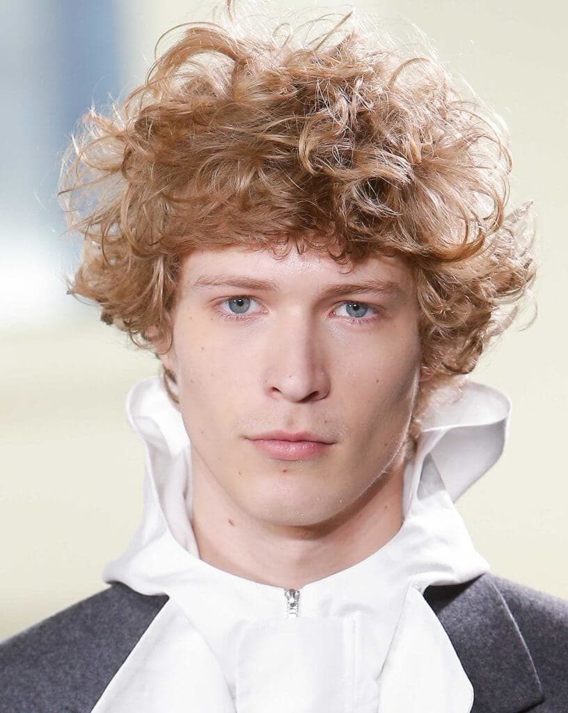 Curly hairstyles for men: Man on runway with blonde curly messy hair wearing a high neck ruffle shirt.