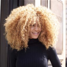 Mulher posa com cabelo afro pintado de loiro e ilustra Como usar modelador de cachos no cabelo crespo.