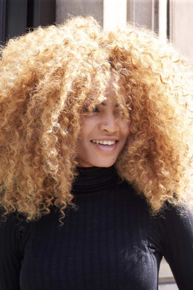 Mulher posa com cabelo afro pintado de loiro e ilustra Como usar modelador de cachos no cabelo crespo.