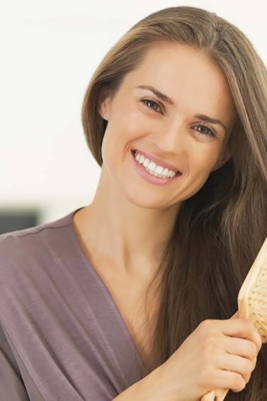 Mulher usa escova raquete enquanto veste roupão lilás e sorri.