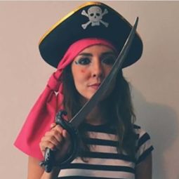 Mulher usa chapéu de pirata, com lenço rosa no cabelo de outros carnavais e camiseta listrada