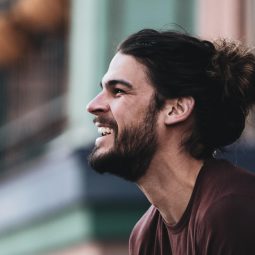 Homem de cabelo longo preso e camiseta marrom sorri ao ar livre