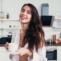 mulher com cabelo longo na cozinha tomando café e sorrindo
