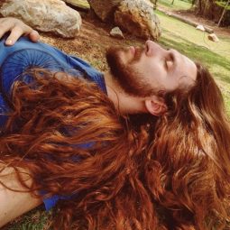 homem de cabelo comprido e acobreado deita na grama de olhos fechados