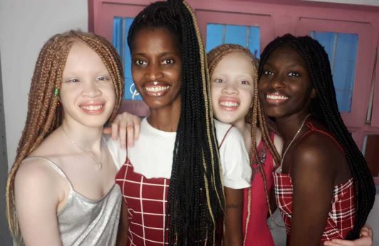 Mãe e filhas sorriem, abraçadas. Elas são negras e usam tranças em seus cabelos e duas das filhas são albinas