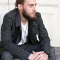 a bearded man with long hair bun sitting on the stair wearing black jacket and white t-shirt