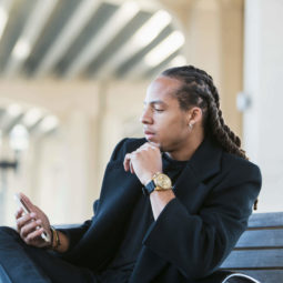a man with long dreadlock hair sitting outside checking his phone wearing a black coat