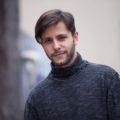 a bearded man with short side bang hair smiling and standing outside wearing a grey sweater