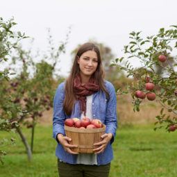 a woman with long brown hair standing in an apple garden after picking up the fruits