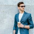 a wavy man standing outside wearing sunglasses and blue suit standing outside while holding a cup of coffee