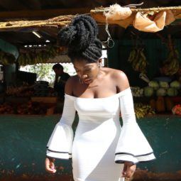 an afro woman with twisted deadlocks hair wearing white dress standing outside near a fruit stalls