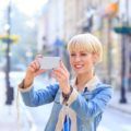 a smiling blonde woman with short bob hair wearing denim outfit taking a picture in a city