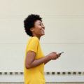 a side view of a black woman with short afro hairstyle wearing yellow tees while playing smartphone outside