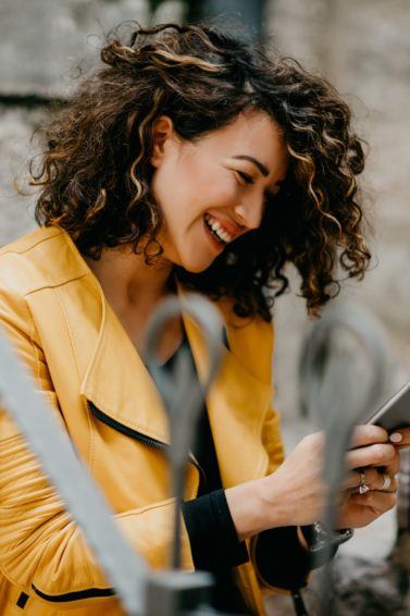 a cheerful curly woman sitting outside playing her phone
