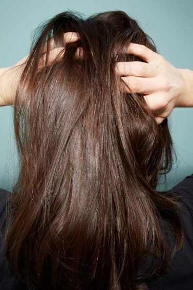 Itchy scalp causes: Back view of a woman with brunette straight hair with her hands in her hair