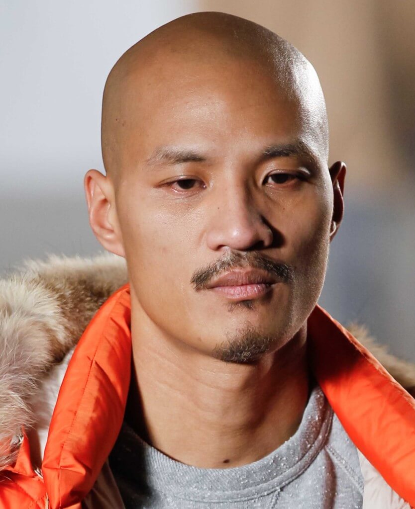 male model on the runway wearing a orange jacket with a fur trim hood with a full shaven head and a moustache