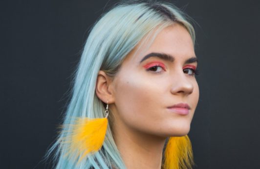 Does dyeing your hair damage it: Woman with sky blue hair, wearing yellow fluffy earrings with furry jacket posing for a street style shot