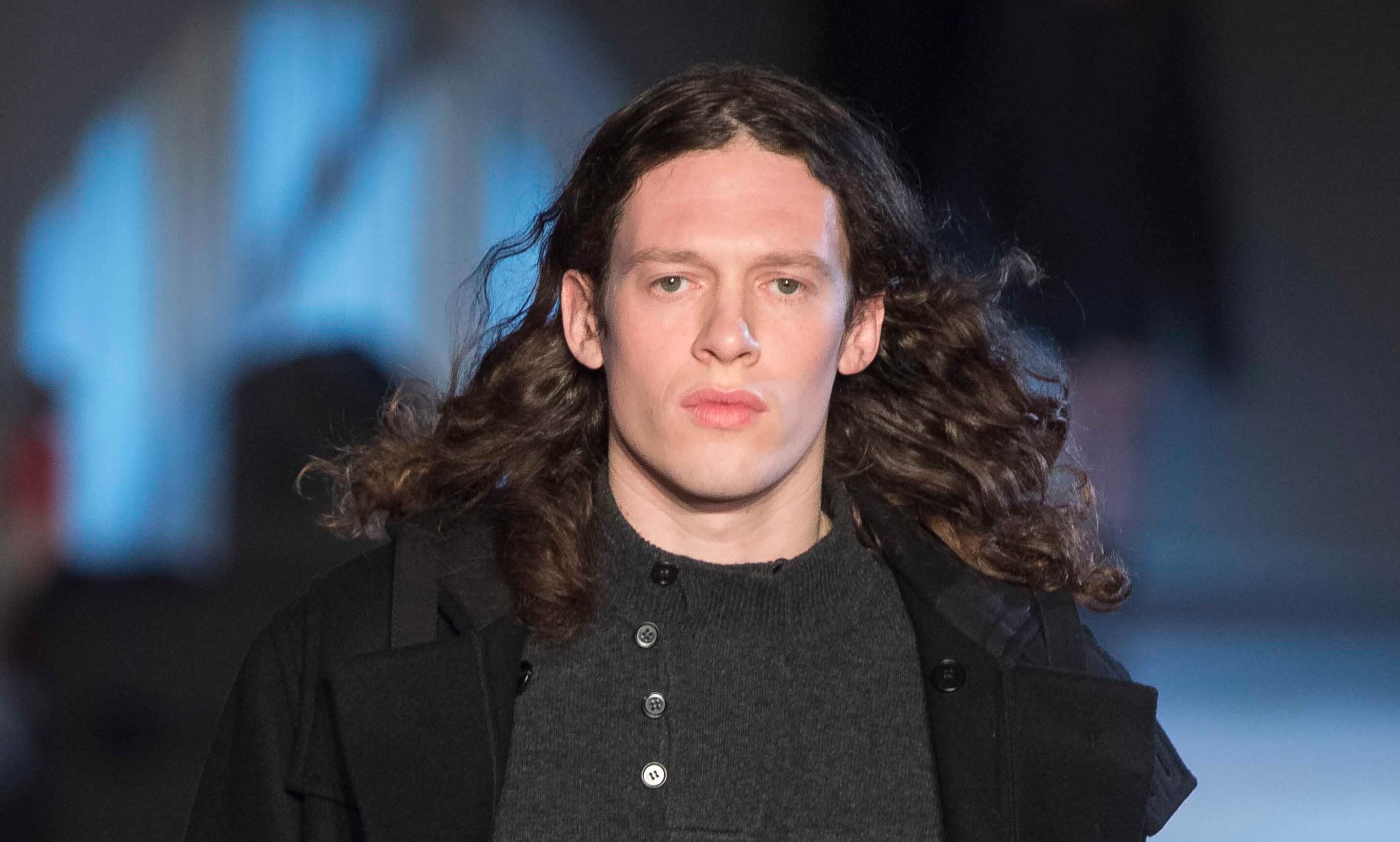 male model on the runway wearing a dark outfit with this dark brown long hair worn loose and in curls