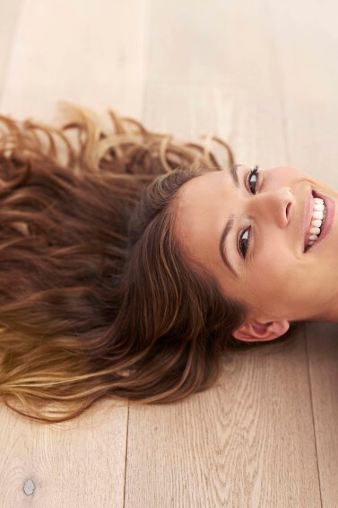 a brunette girl with wavy hair lying down at the floor smiling at the camera