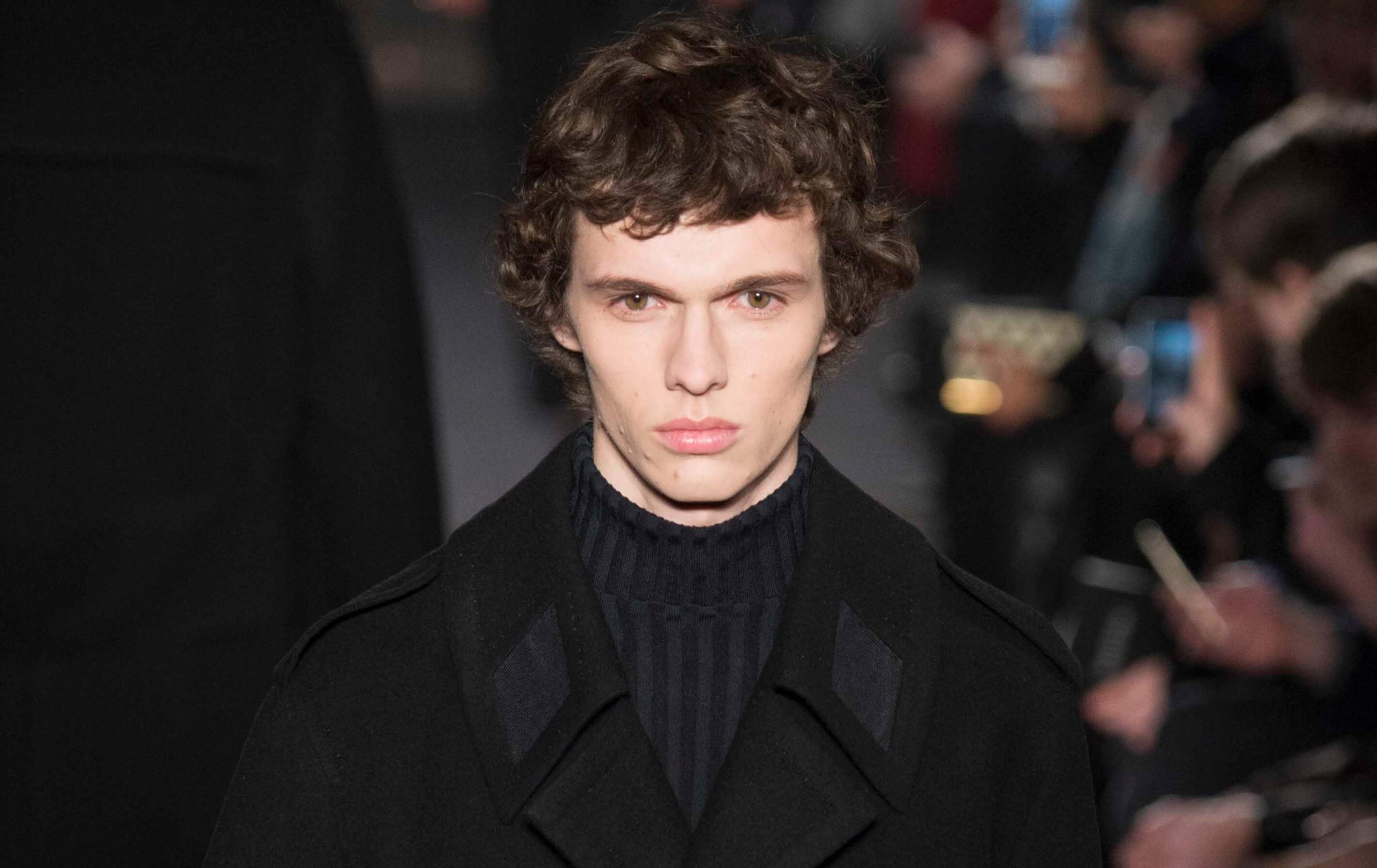male model on the runway wearing a dark outfit with his dark brown hair worn short and in curls