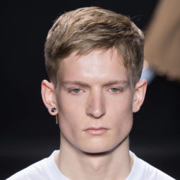 male model on the runway wearing a white top and an earring with his dirty blonde hair worn in a neat crop