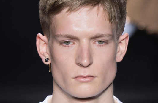 male model on the runway wearing a white top and an earring with his dirty blonde hair worn in a neat crop