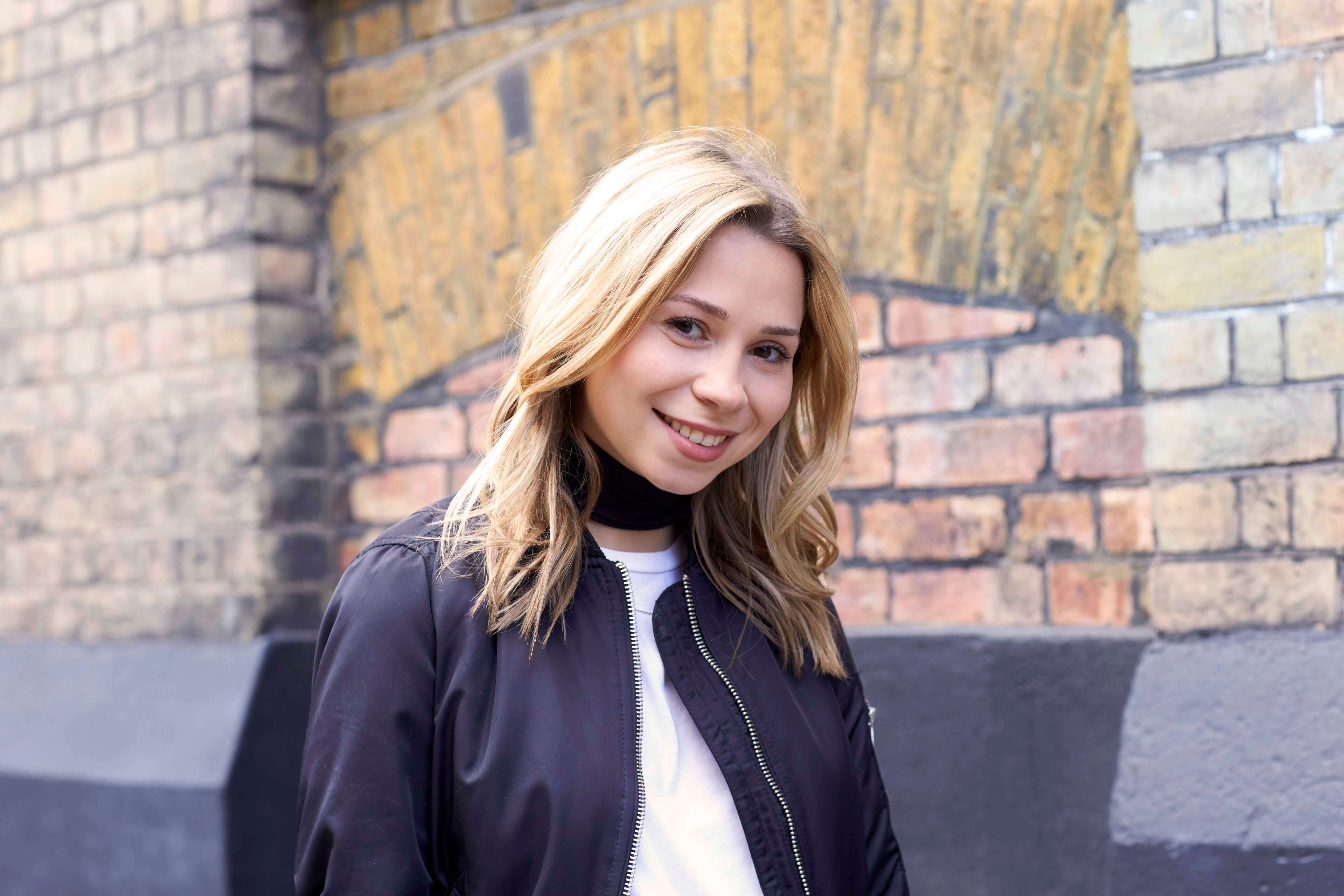 Medium length hairstyles for fine hair: Woman with blonde medium length hair with layers wearing a black bomber jacket and white top.
