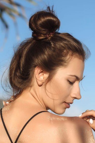 summer hair tips: close up shot of woman on the beach with dark brown hair fashioned into a bun, wearing a bikini top and posing