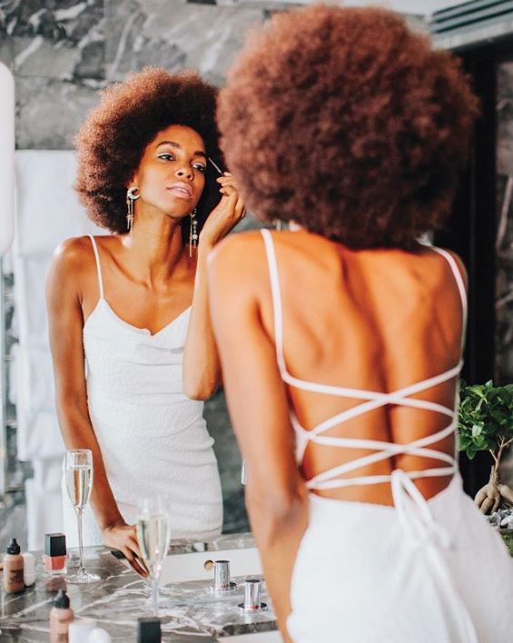Wedding hairstyles for short hair: Woman with large natural afro hair wearing a strappy wedding dress doing makeup in the mirror.