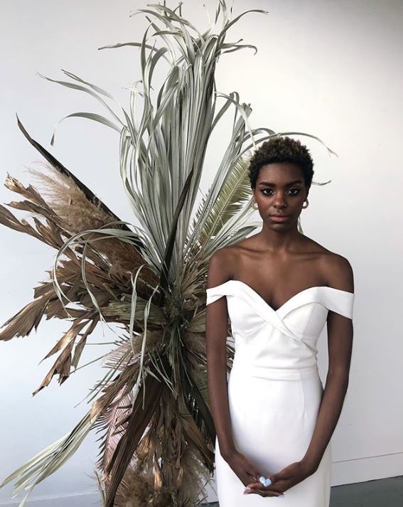 Wedding hairstyles for short hair: Woman with short natural afro hair wearing aoff the shoulder wedding dress standing in front of a large plant