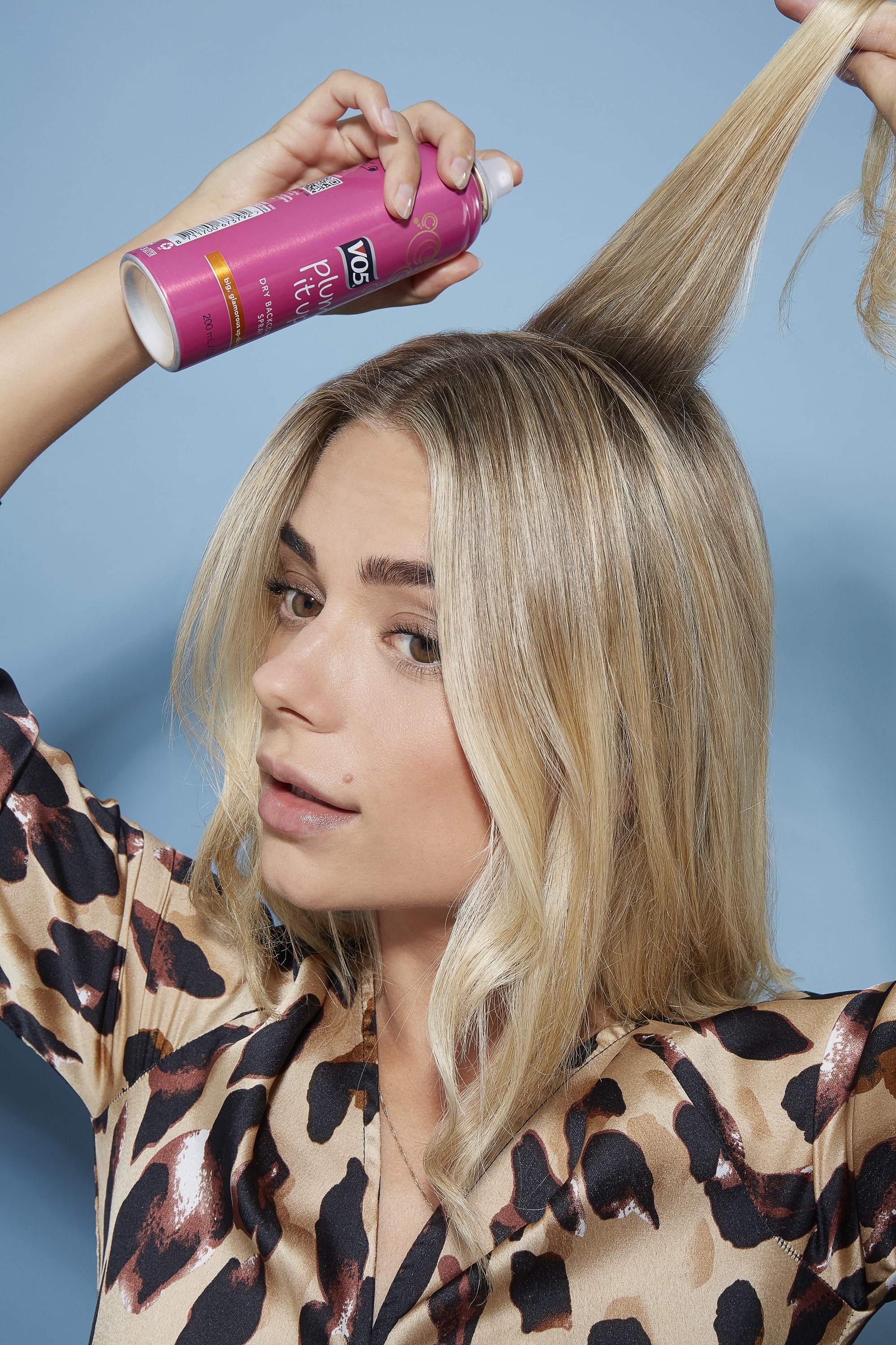 blonde model spraying product into roots of mdeium length straight hair wearing leopard print top
