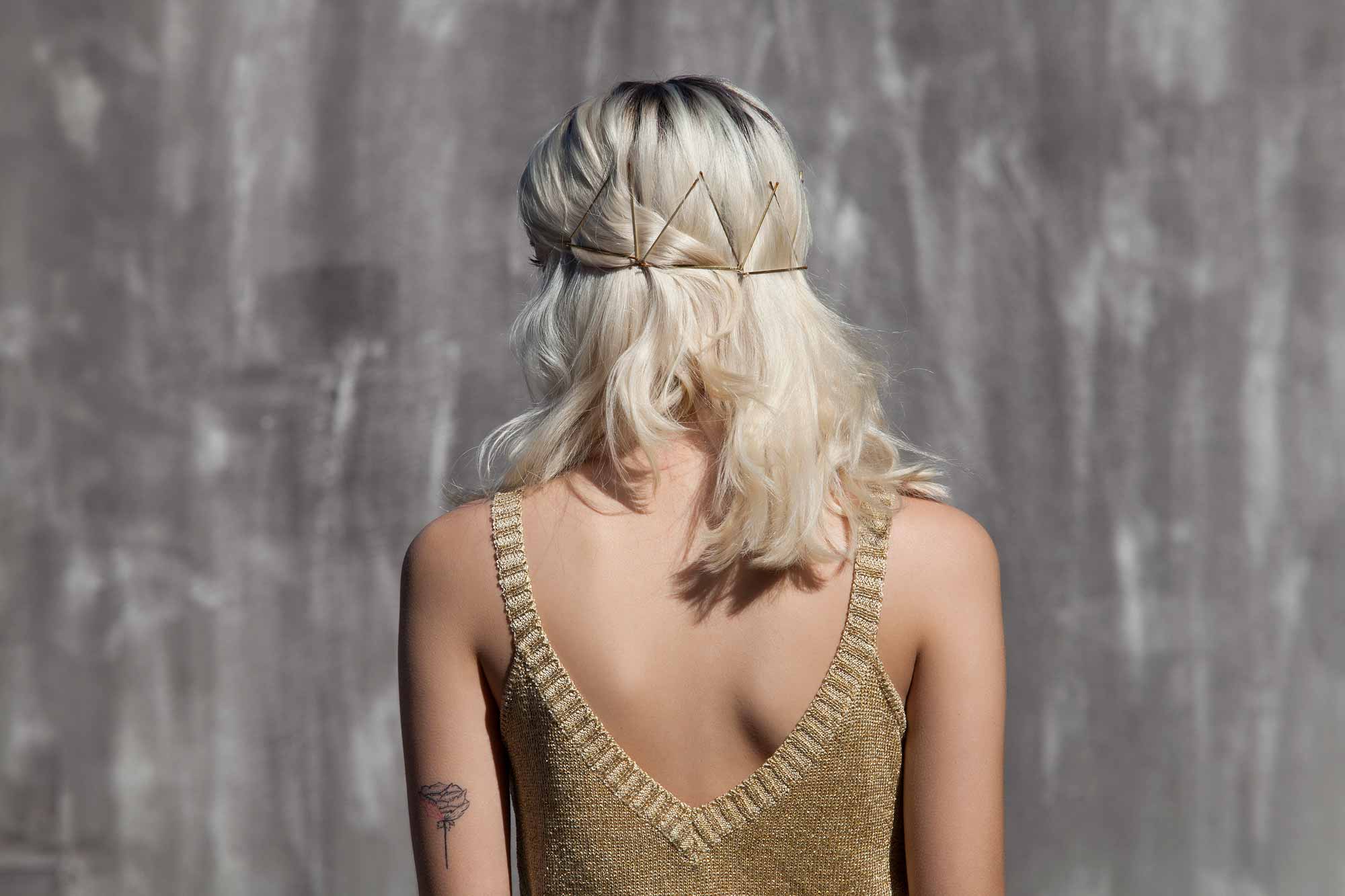 Bobby pin hairstyles: Close up backshot of a woman with messy bleach blonde medium hair styled with a triangle bobby pins, wearing a gold top and posing outside