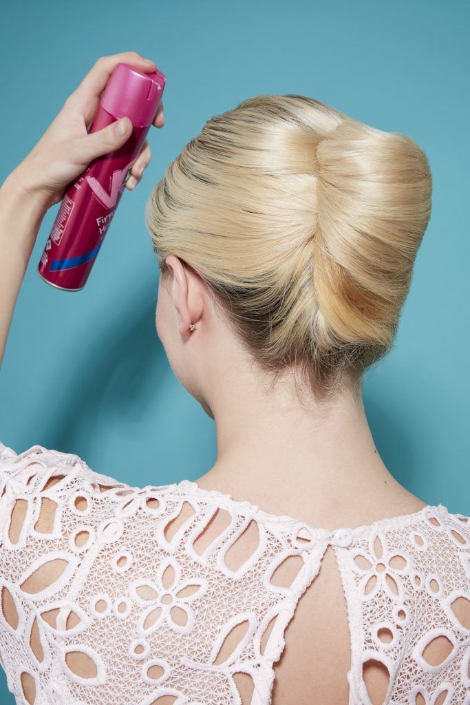 model with light blonde hair rolled into a french twist updo, spraying hairspray to hold it in place