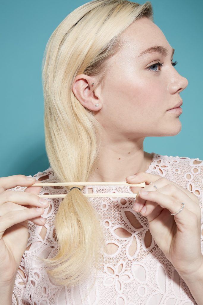 model with light blonde hair and darker roots using chopsticks hold her hair ready to roll it into a an updo