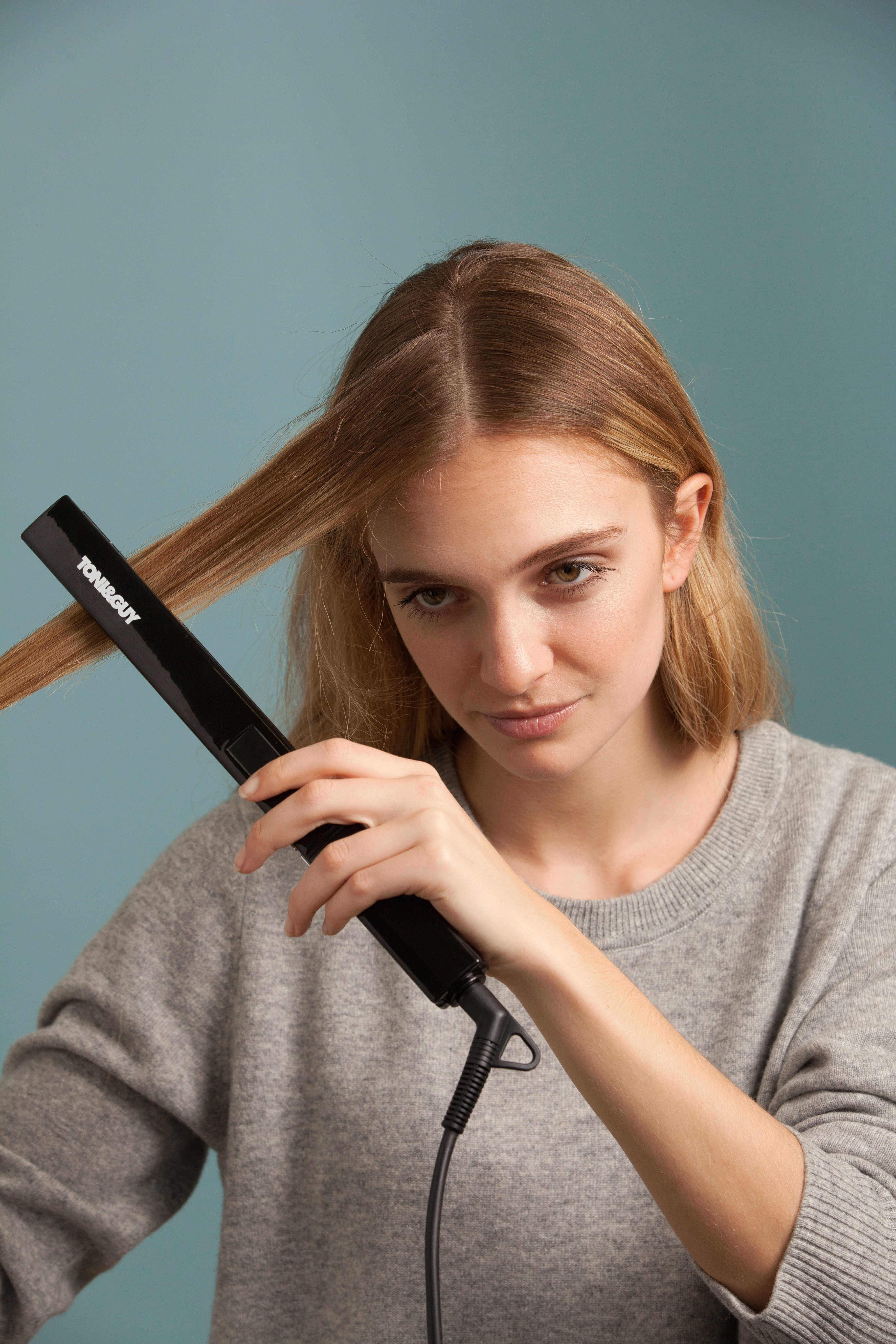 Hair straightening treatment: Blonde model with straight hair using a hair straightener wearing a grey jumper.
