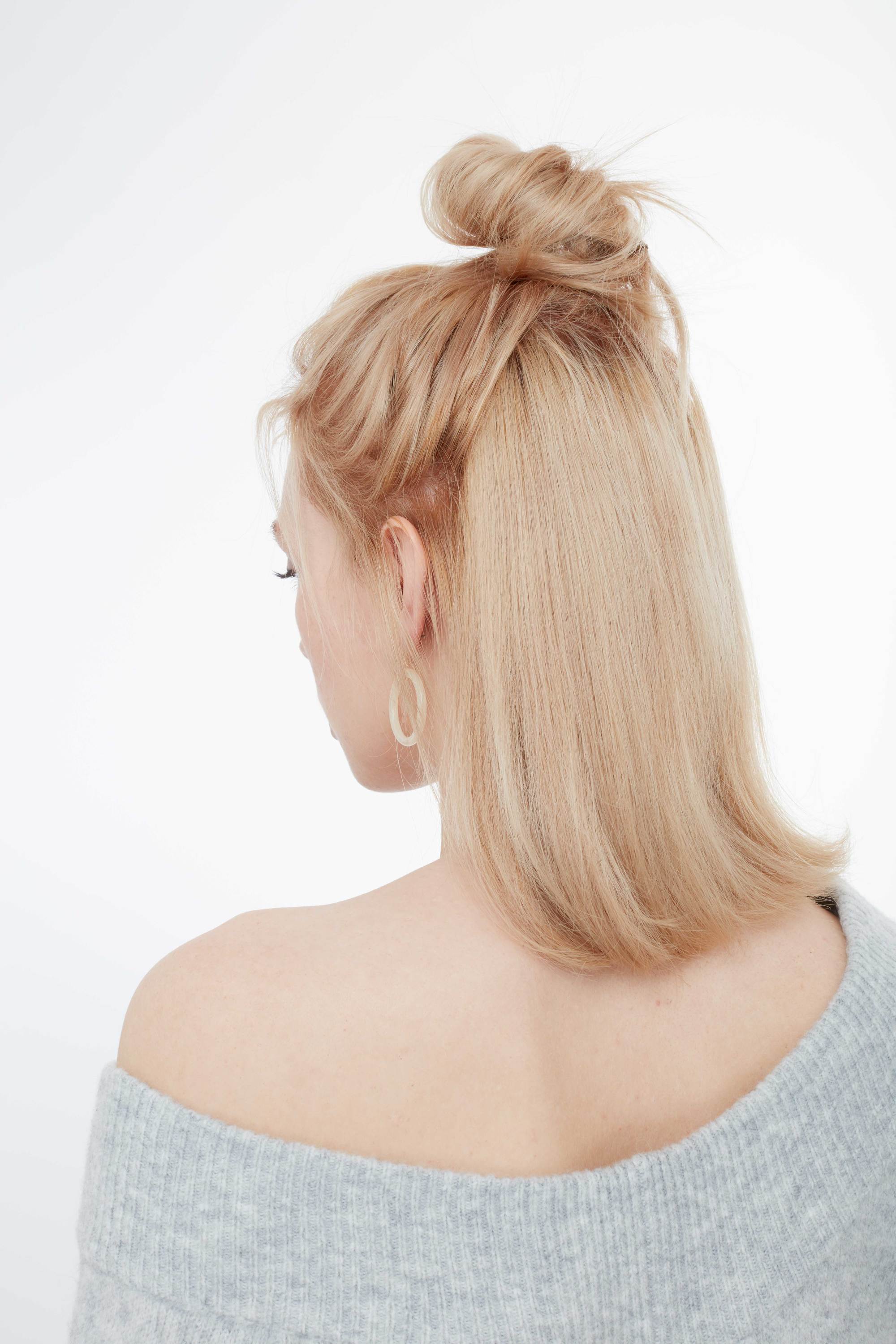 Back shot of woman with golden blonde thin hair styled into a half-up, half-down bun hairstyle,m wearing grey and posing in a the Al Things Hair studio