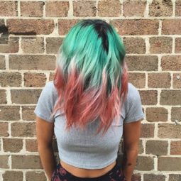 close up shot of woman with watermelon hair, wearing grey top and black shorts and standing against a brick wall