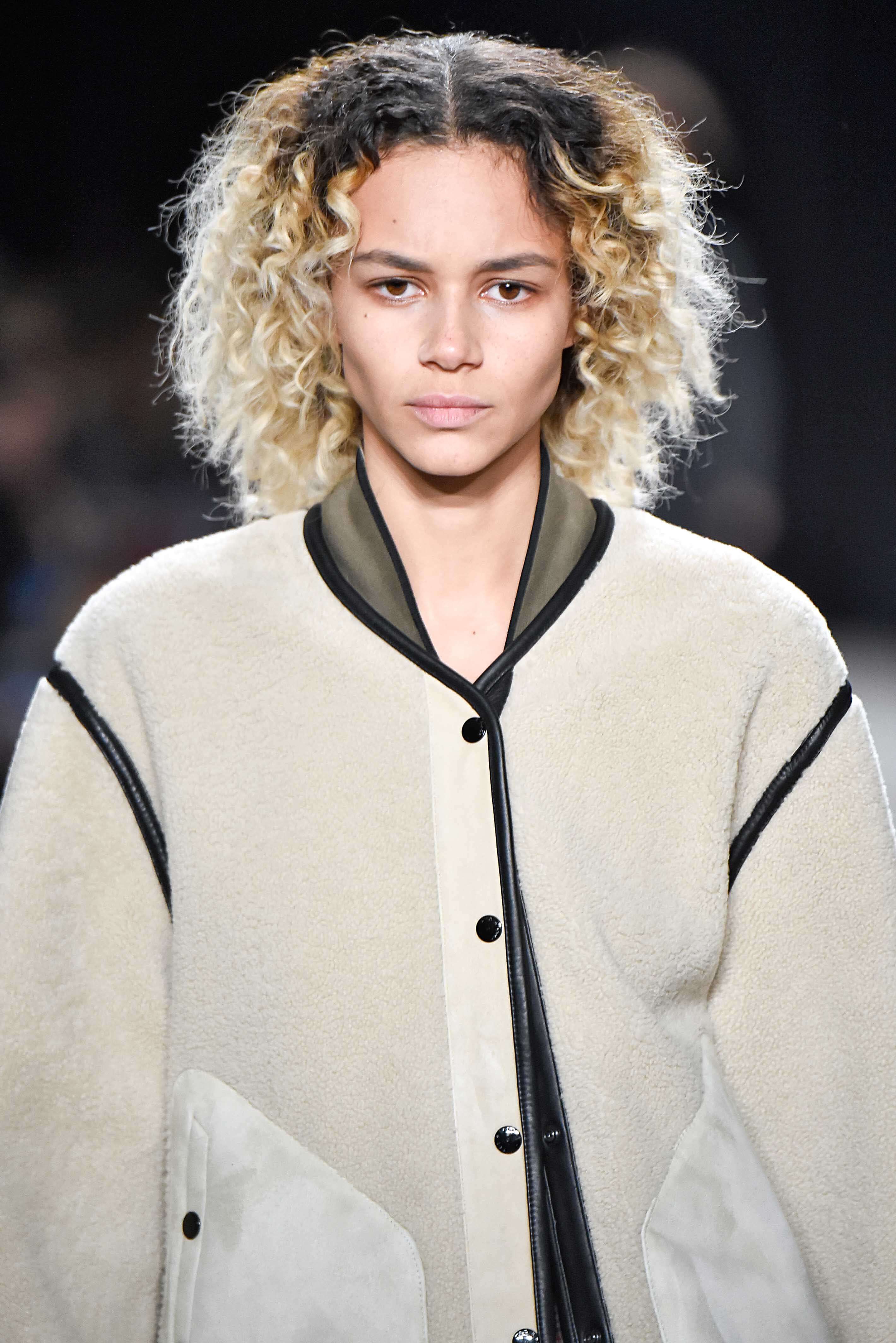 Shades of blonde hair: Woman with platinum blonde naturally curly hair with dark brown roots on runway wearing a bomber jacket.