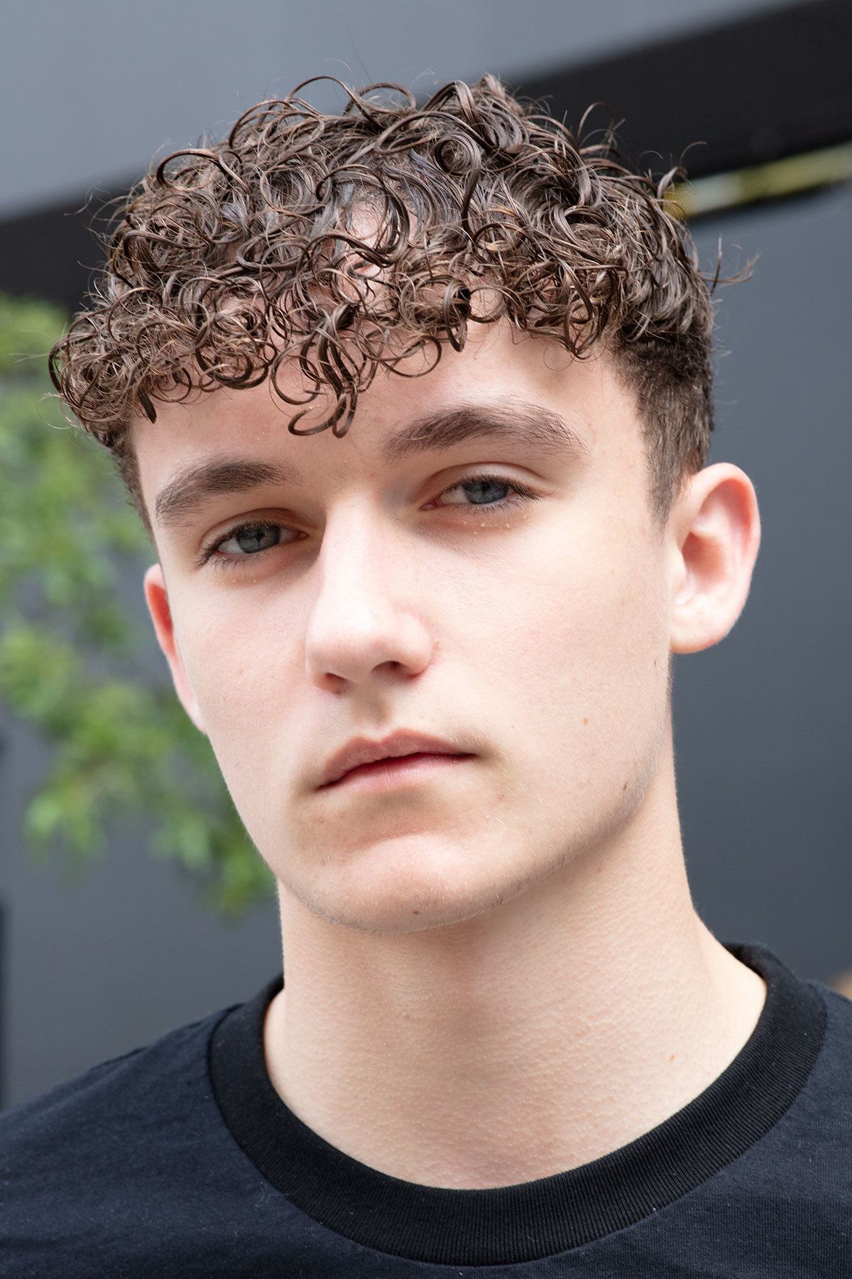 Best hair styling products for men: Close up shot of a man with chocolate brown curly French crop, wearing a black shirt and posing on the street.