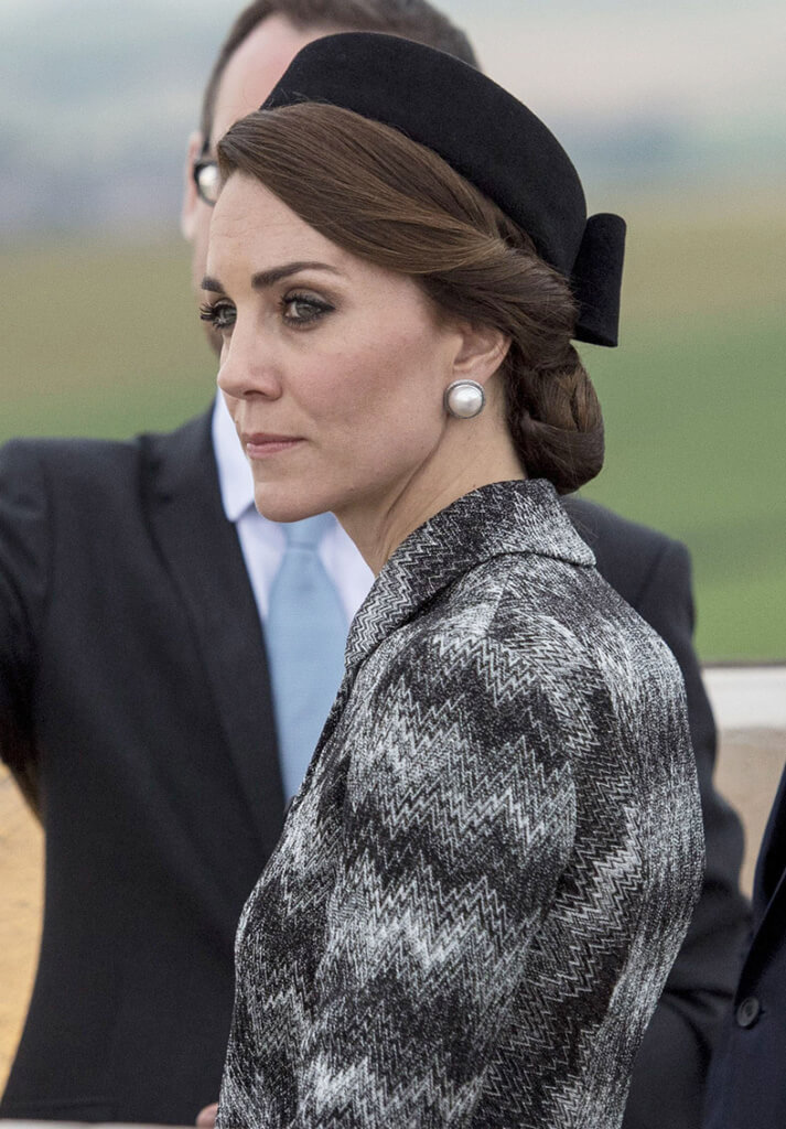 kate middleton wearing a grey outfit with her hair twisted into a low bun with a hairnet accessory and a black hat