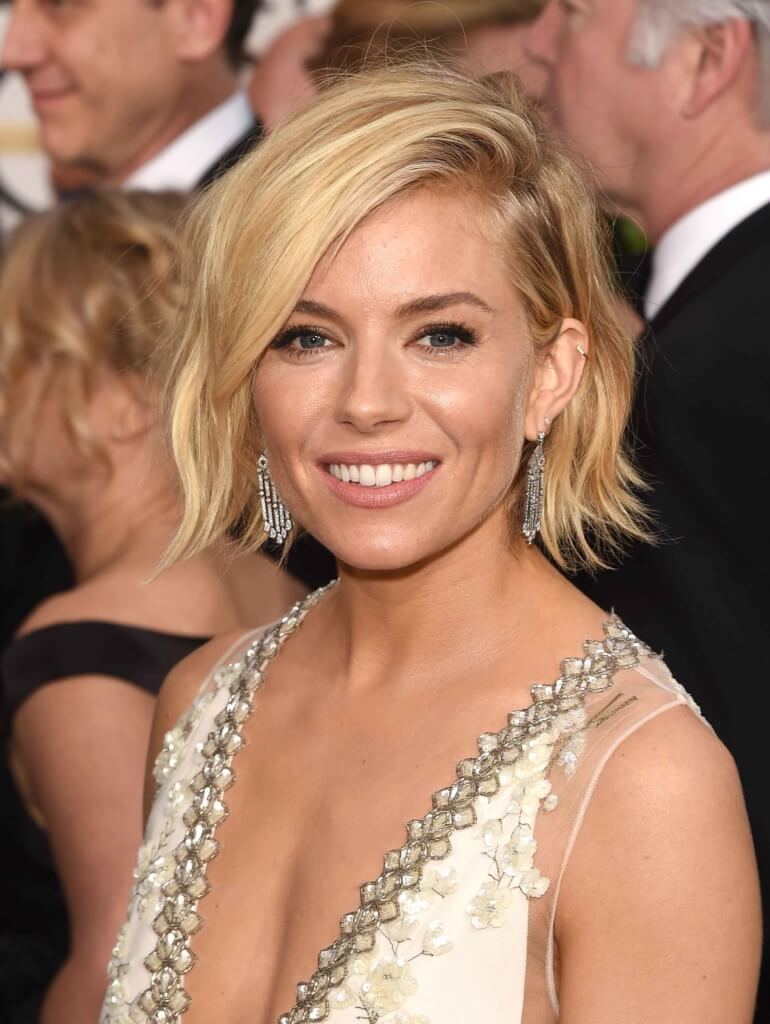 wavy blonde bob on Sienna Miller wearing a white and silver dress with droplet earrings in the red carpet